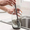 Hands are using a silicone bottle brush with a long handle to clean inside a glass container.