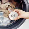 A hand is holding a light beige wool dryer ball inside a dark laundry basket.