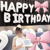 A person is holding a pink birthday cake in front of a white and pink banner decoration set.