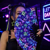A woman holds a holographic gradient rave hand fan with purple and blue floral patterns in a dark, neon-lit setting.