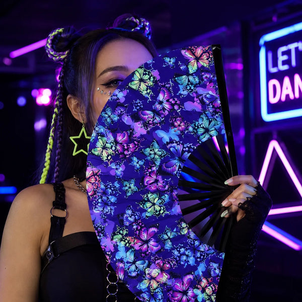 A woman holds a holographic gradient rave hand fan with purple and blue floral patterns in a dark, neon-lit setting.