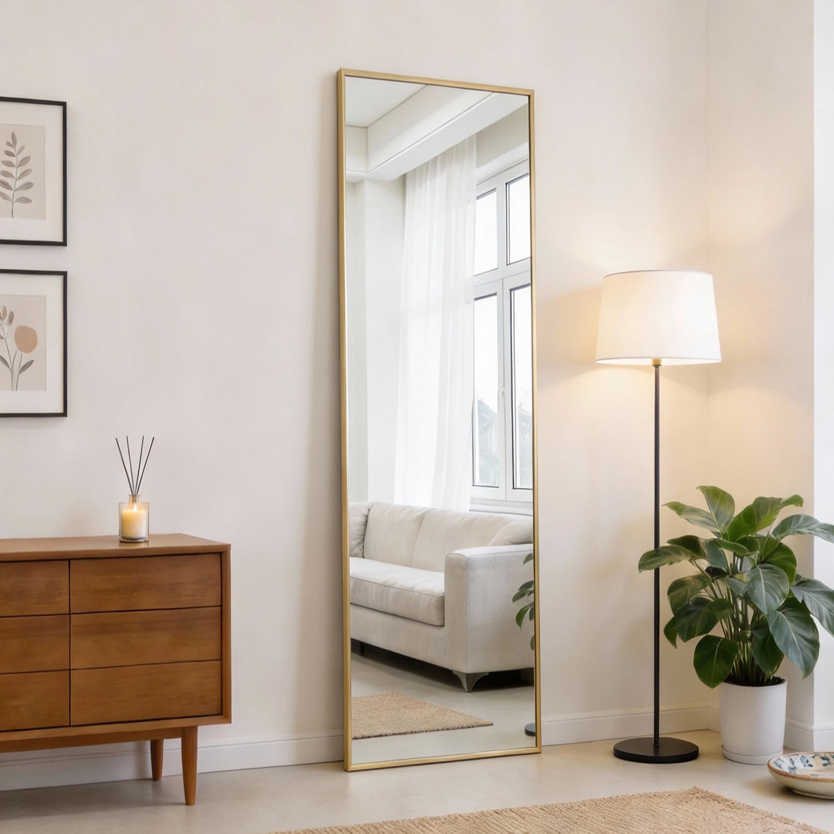A tall full body wall mirror with a gold frame reflecting a bright room and window.