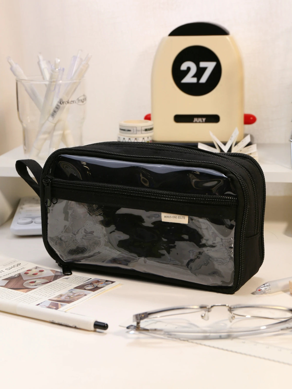 A black transparent pencil pouch is displayed on a white surface next to a yellow and black numbered object.