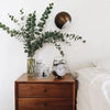 A dark bronze lamp illuminates a glass vase holding green foliage and a heart-shaped acrylic plaque on a wooden nightstand.
