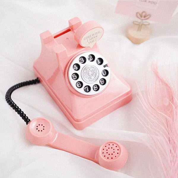 A pink retro phone money jar with a coiled cord and a decorative flower is shown on white fabric.