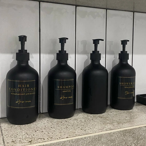 Three black refillable soap dispenser bottles labeled Hair Conditioner, Shampoo, and Shower Gel are displayed on a counter.