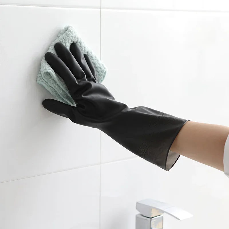 A hand wearing a black rubber cleaning glove is wiping a white tile with a green cleaning cloth.