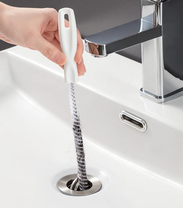 A hand is holding a flexible drain cleaning brush with a white handle, positioned over a sink drain.
