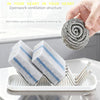 A hand holds a coiled gray cleaning sponge, with two blue-striped sponges resting in a stainless steel rack on a white tray.