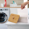 A hand is placing a bamboo-lidded dryer sheet container on a white countertop next to a washing machine.