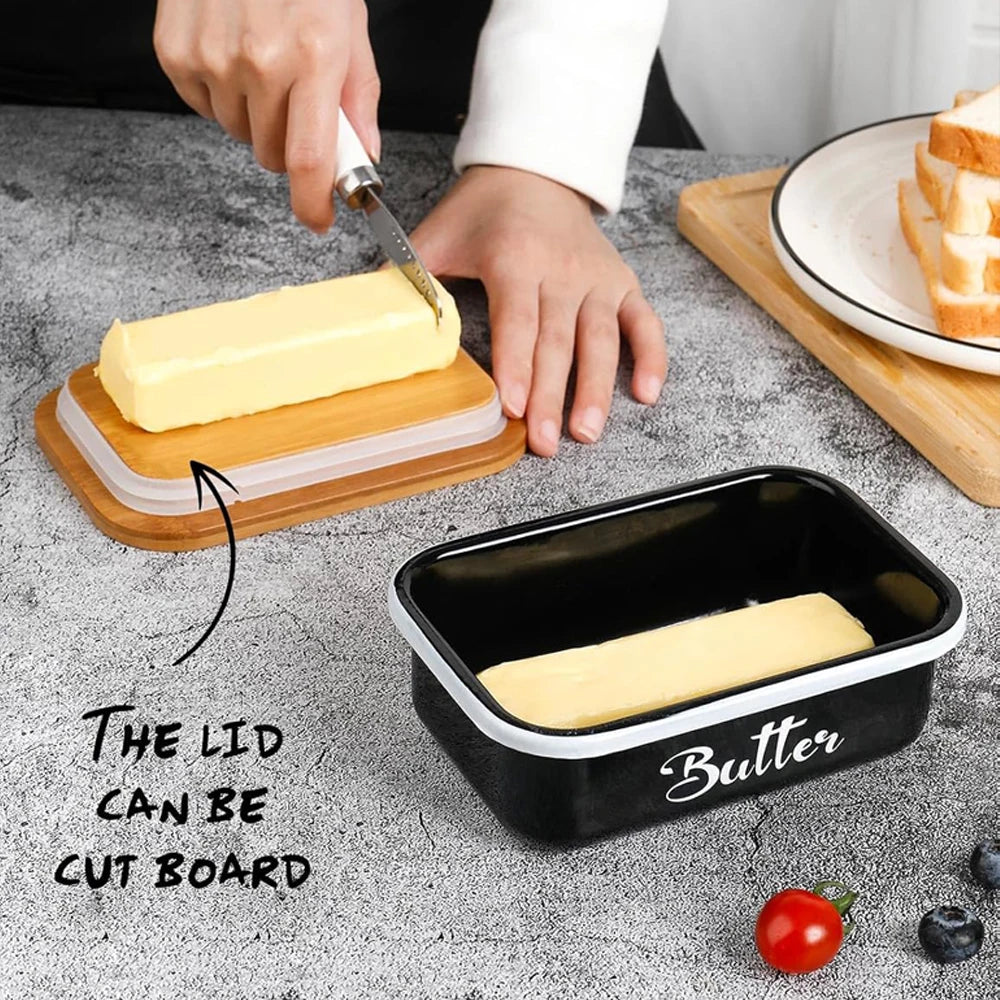 Une main coupe un bloc de beurre avec un couteau sur une planche en bois, à côté d'un élégant pot à beurre en métal.