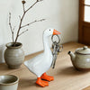A white goose key chain sculpture with orange beak and feet resting on a wooden table next to a ceramic teapot and a bare bra