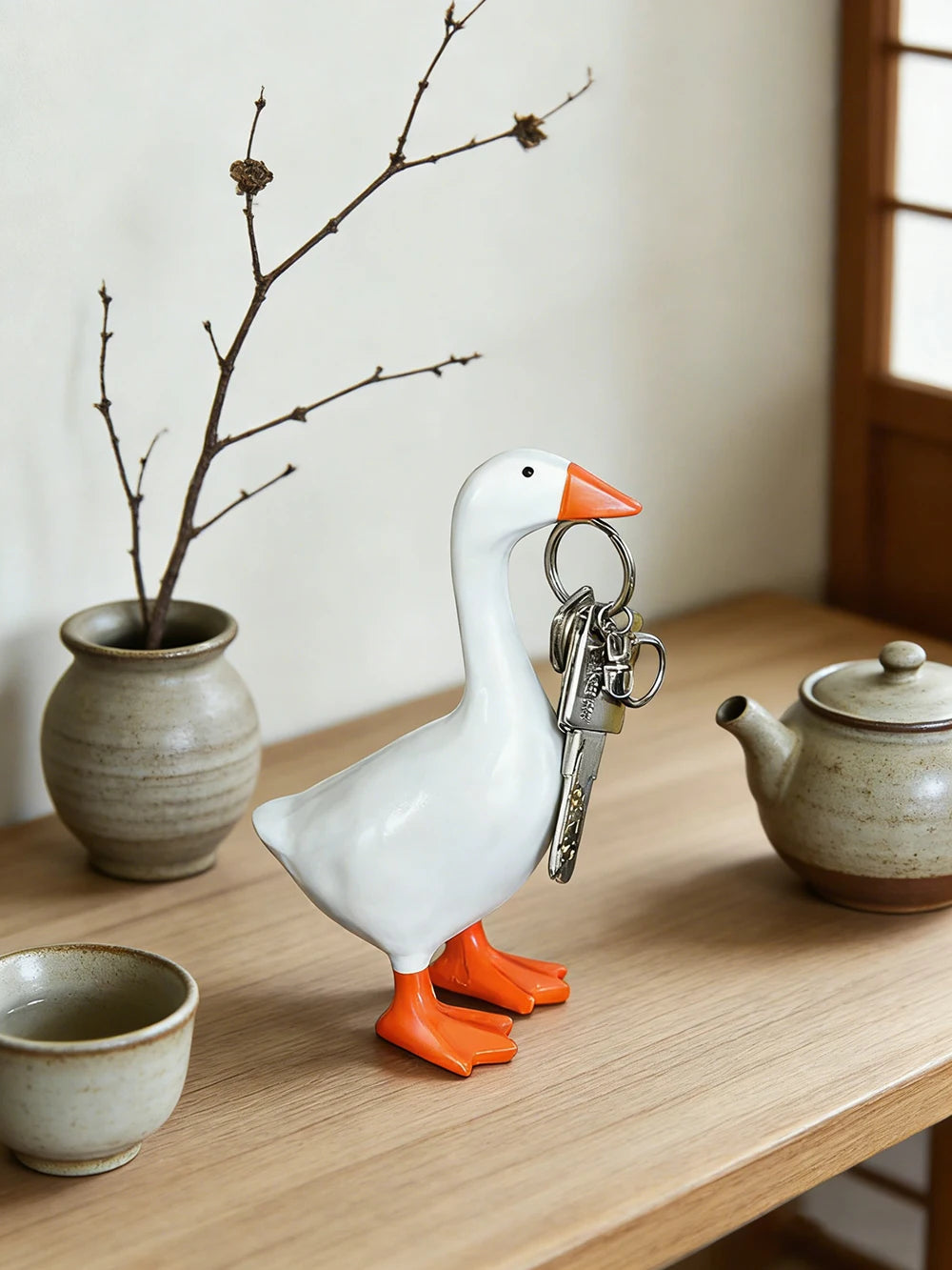 A white goose key chain sculpture with orange beak and feet resting on a wooden table next to a ceramic teapot and a bare bra