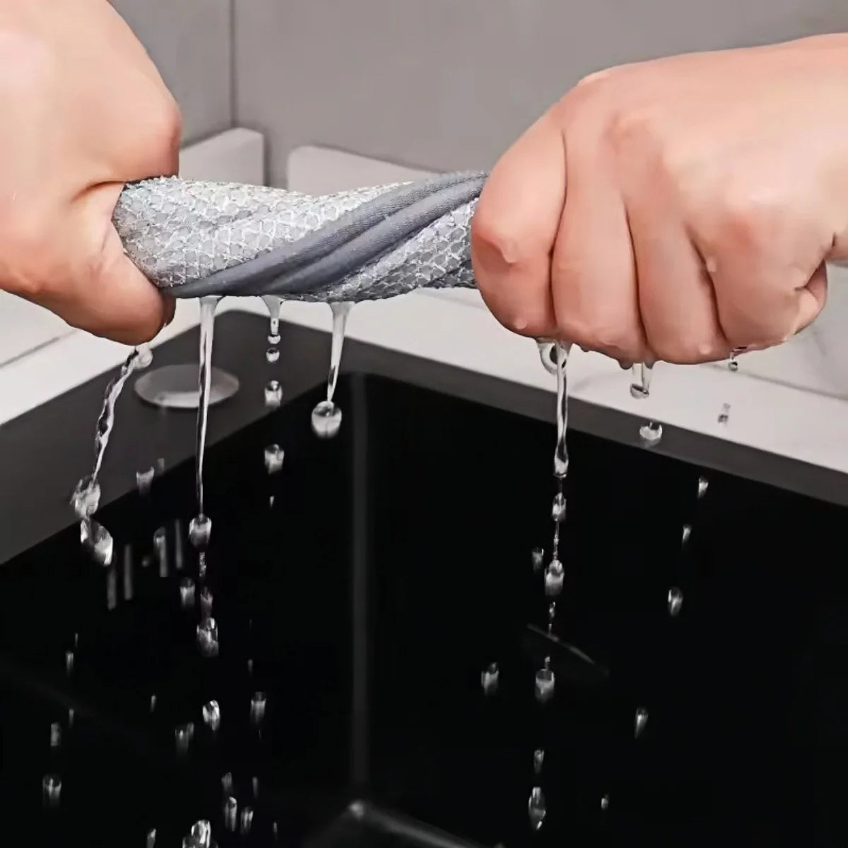 Hände tropfen Wasser in eine schwarze Spüle, wobei ein graues Reinigungstuch zum Waschen verwendet wird.
