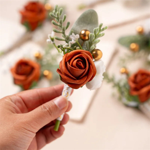 A hand holds a vintage orange silk boutonniere featuring roses, greenery, and gold accents.