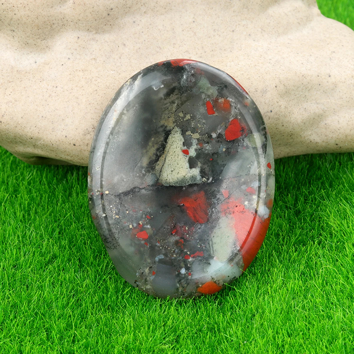 A polished, oval natural quartz thumb stone with black and red inclusions rests on bright green grass.