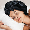 A woman wearing a black silky satin sleep bonnet rests peacefully on white bedding.