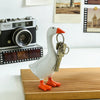 A white goose sculpture with orange feet holds a brass key chain.