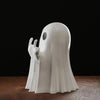 A white ghost statue decoration with outstretched hands is displayed on a dark brown surface against a black background.