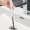 A hand is holding a white flexible drain cleaning brush with a metal handle over a kitchen sink.