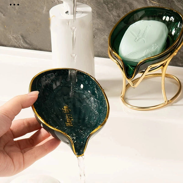 A hand pours water into a decorative leaf-shaped soap dish made of dark green ceramic.