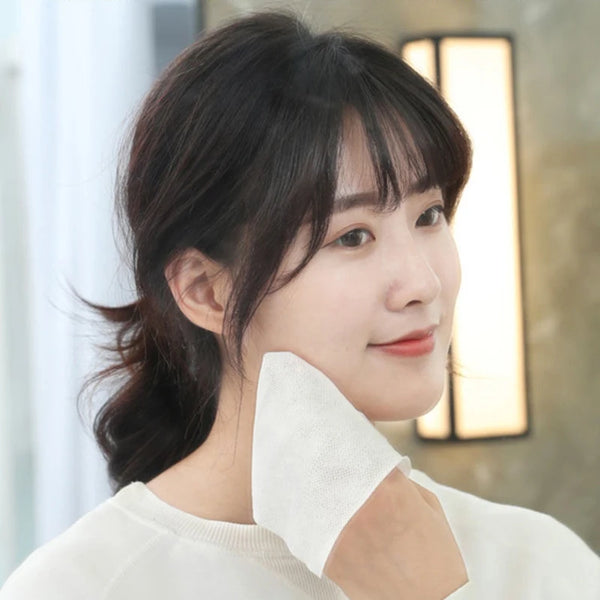 A woman is holding a white disposable cotton face towel near her face in a soft indoor setting.
