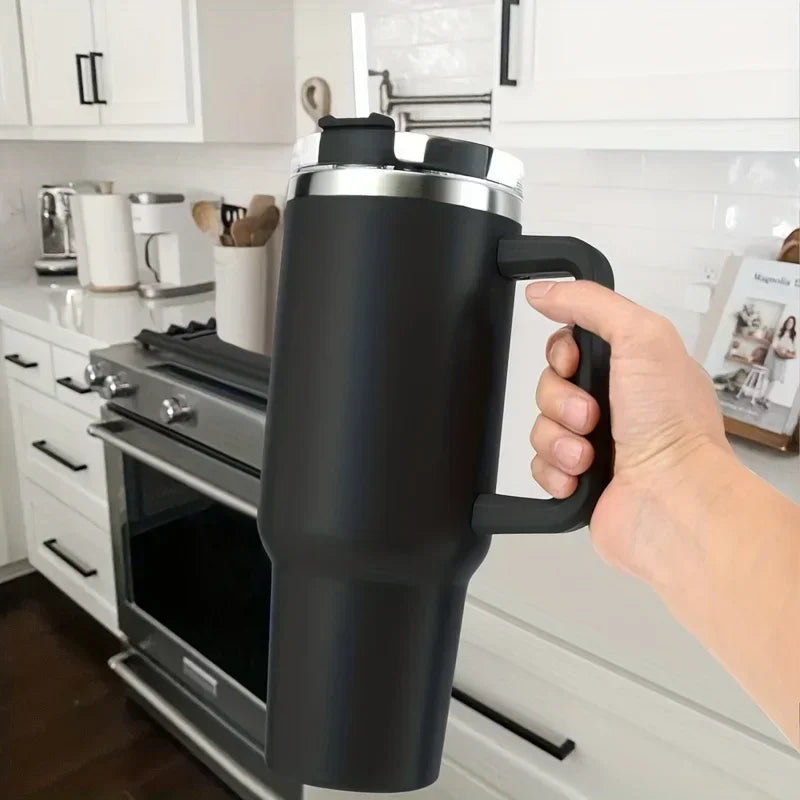 A hand holds a black insulated stainless steel tumbler with a handle and straw in a kitchen setting.