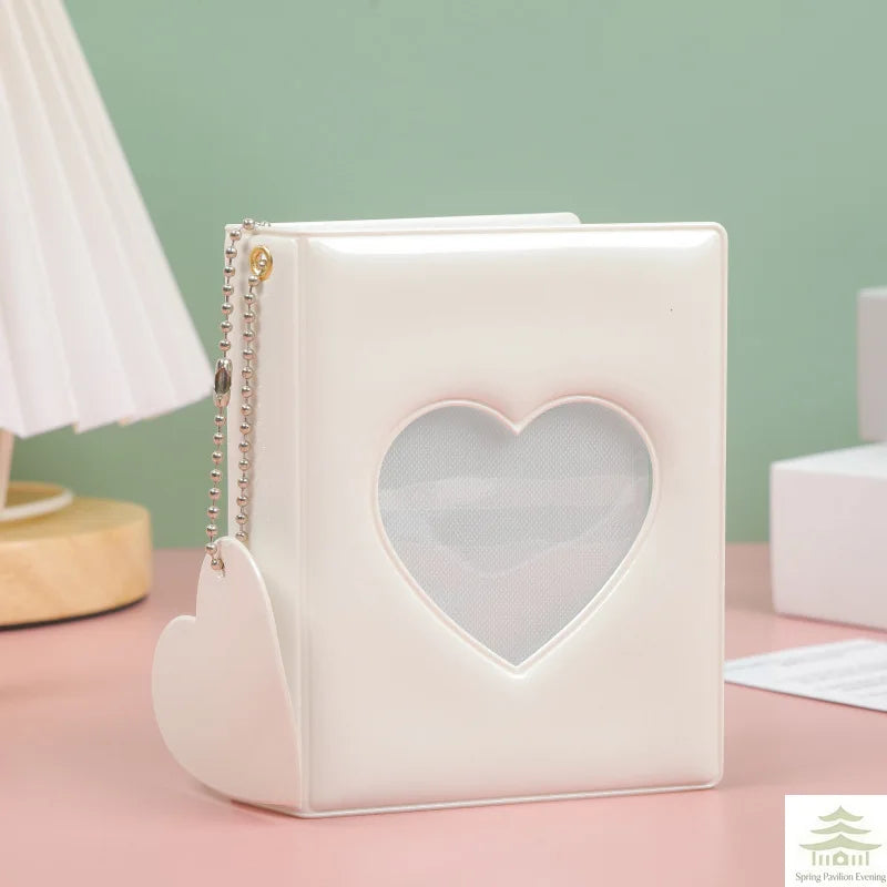 A white mini photo album with a heart-shaped cutout and a chain strap rests on a pink surface.