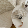 A white embroidered sun umbrella is displayed on a round white plate next to a pillow.