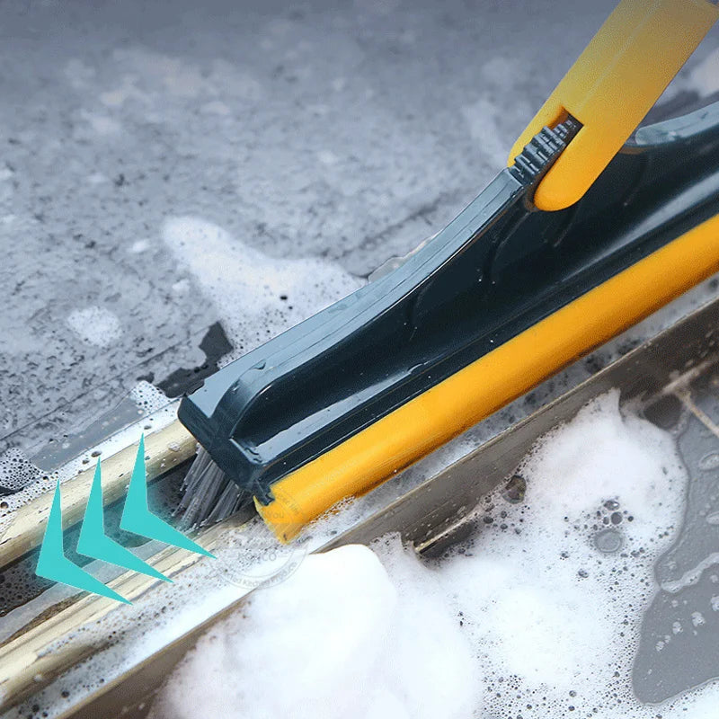 A yellow and black 2-in-1 floor scrub brush is cleaning soapy water off a wooden surface.