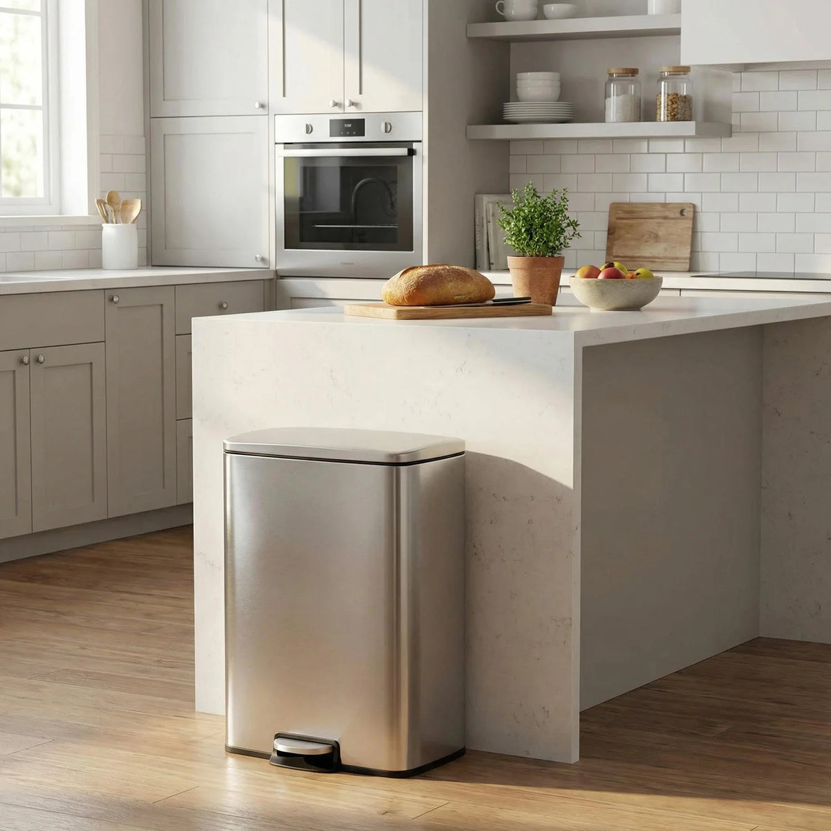 A stainless steel pedal trash bin is placed on a kitchen counter in a modern, light-colored room.