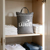 A gray laundry bag filled with white towels is displayed on a wooden shelf inside a cabinet.