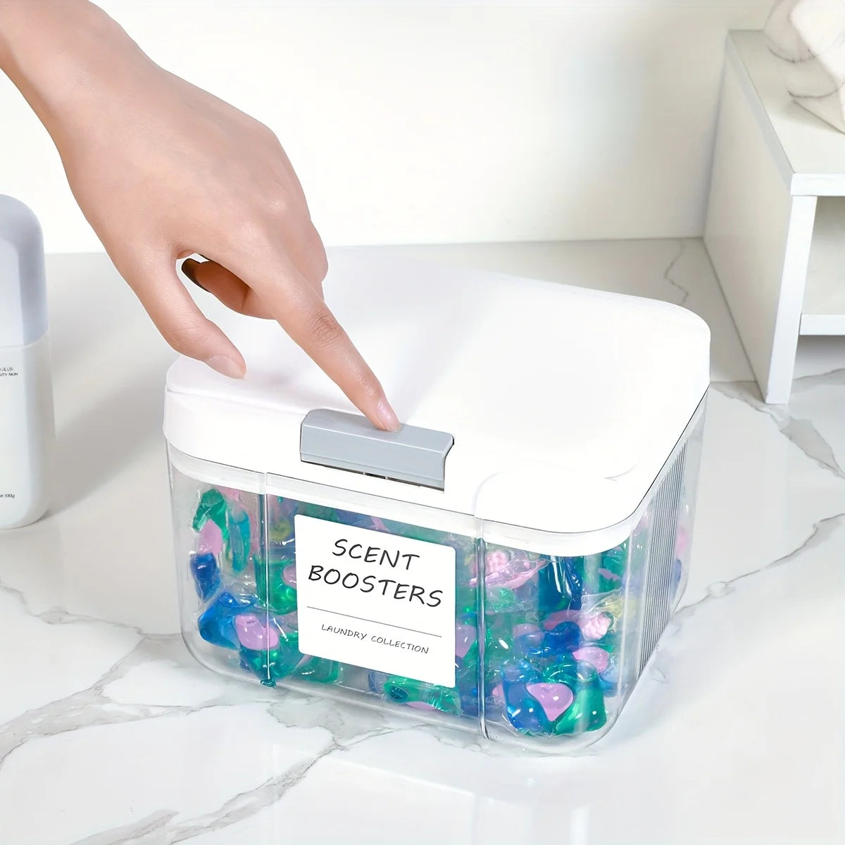 A hand places a silver dispenser onto a clear plastic laundry pod organizer filled with colorful laundry boosters.