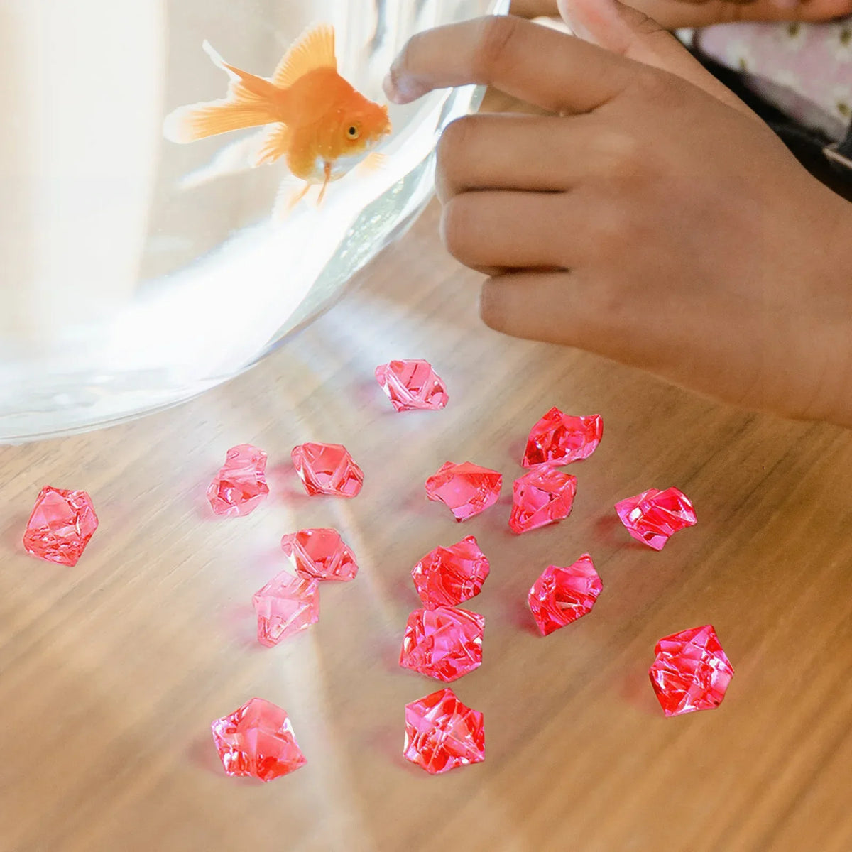 A hand is holding a small orange fish near crushed pink and red crystal ice stones on a wooden surface.