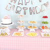A pink birthday party table decorated with a banner, tiered cakes, and various fruits and candies on stands.