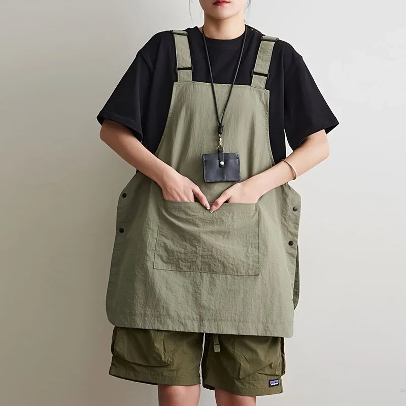 A woman wearing a olive green waterproof outdoor work apron over a black shirt and khaki shorts.