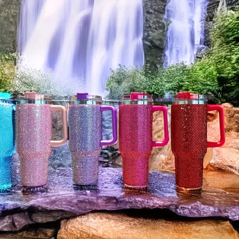 Tres vasos de acero inoxidable con pedrería rosa y roja exhibidos sobre rocas con un fondo de cascadas y vegetación.