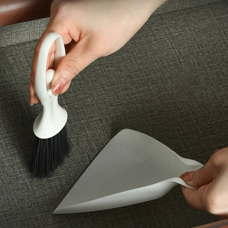 A hand is holding a white mini broom and dustpan set on a dark surface.