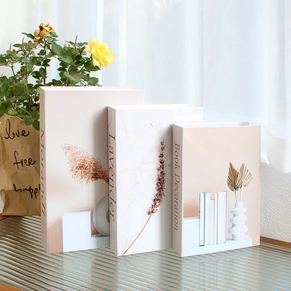 A set of decorative fake books in pale pink and white with dried floral accents displayed on a glass surface.