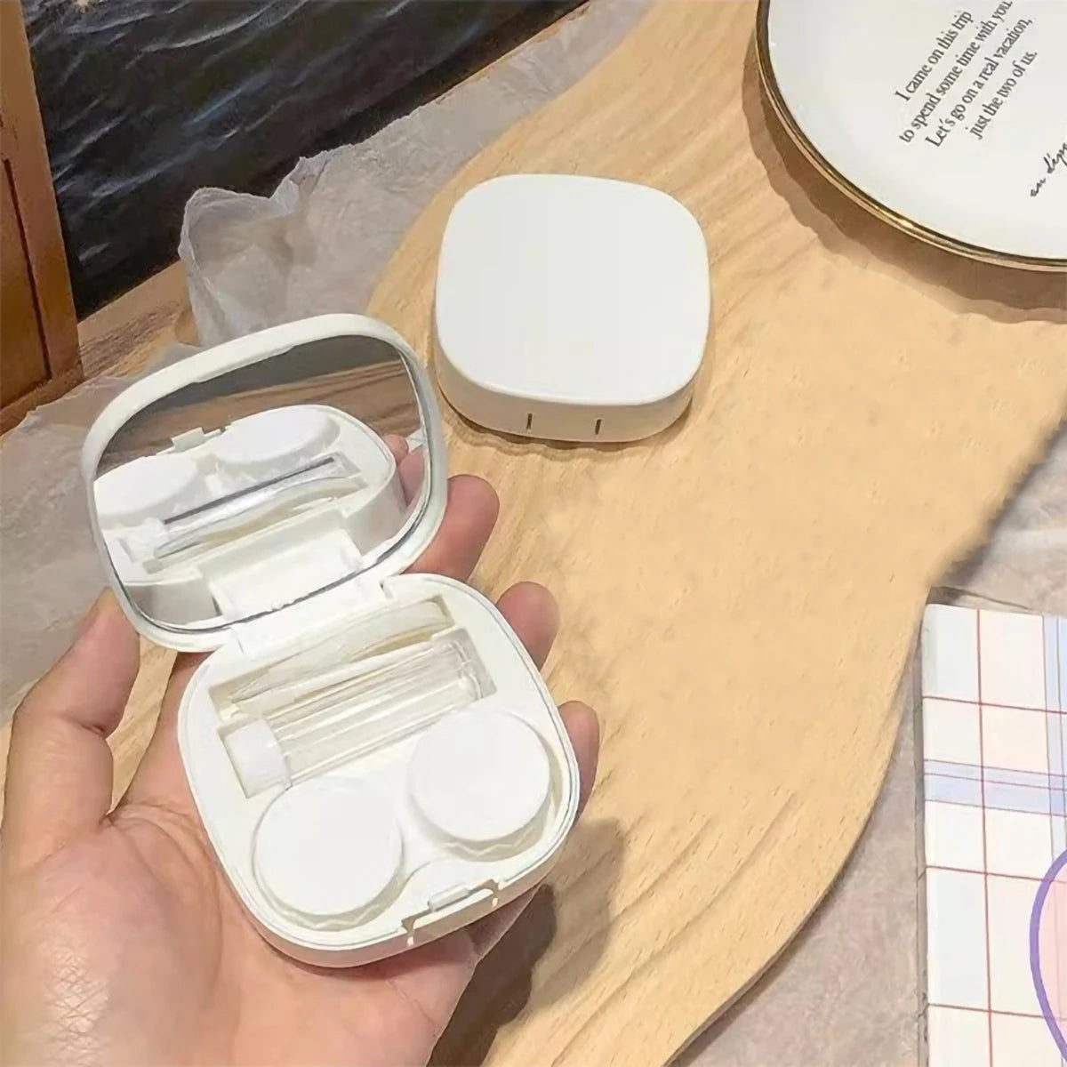 Eine Hand hält eine offene weiße Kontaktlinsenaufbewahrungsbox und eine separate weiße Aufbewahrungskiste auf einer hellen Ho