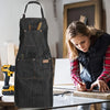 A dark gray denim tool apron with multiple pockets and a handle is shown on a wooden table.