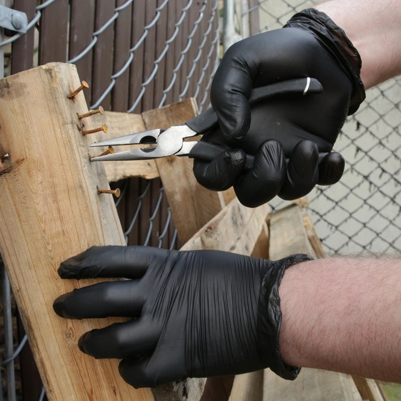 A hand wearing a black disposable nitrile work glove is holding silver pliers near a wooden structure.