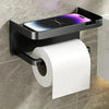 A black no-drill toilet paper holder with a storage shelf mounted on a wall, holding a roll of white paper.
