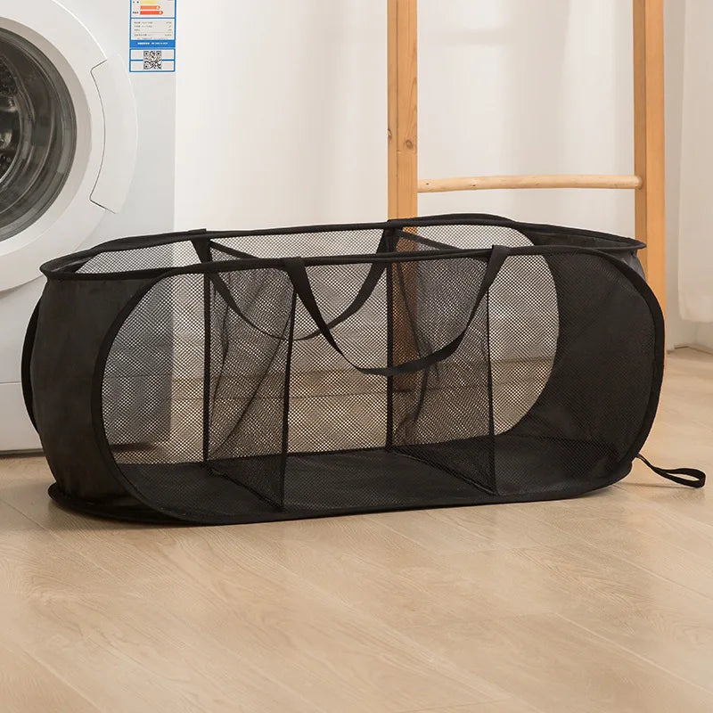 A black foldable mesh laundry basket with a handle is shown on a light wooden floor.