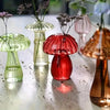 A collection of glass mushroom-shaped vases in various shades of green, pink, and red displayed on a reflective surface.