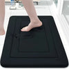 A black non-slip absorbent bath mat is placed on a white tiled floor, with a person's foot resting on the mat.