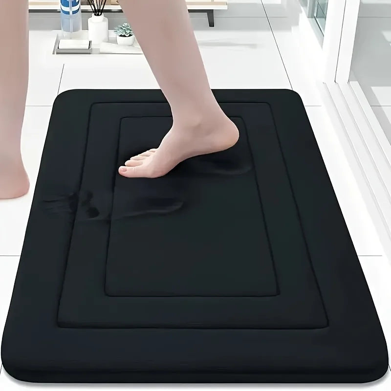 A black non-slip absorbent bath mat is placed on a white tiled floor, with a person's foot resting on the mat.