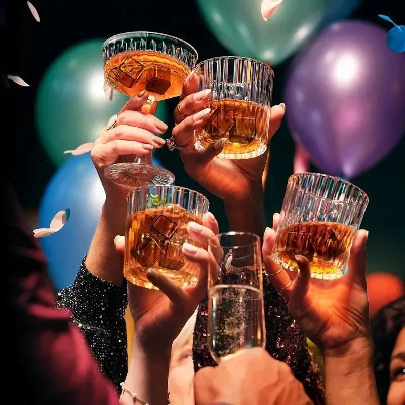 Multiple hands hold various glasses filled with amber whiskey and ice cubes against a blurred background of balloons.