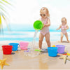 A toddler is playing with a green plastic water splashing bucket on a sandy beach with the ocean in the background.