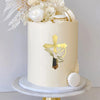 A round cream-colored cake decorated with a gold acrylic cross topper and white floral accents.